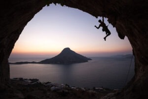 Silhouette of male rock climber climbing cave roof with sea view at sunset