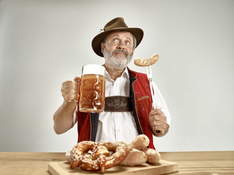 Older happy smiling man with beer dressed in traditional Austrian or Bavarian costume holding mug of beer at pub or studio.
