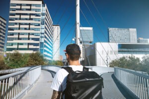 Hipster male with a large black backpack travels on a diverging bridge against city landscape and bright blue sky